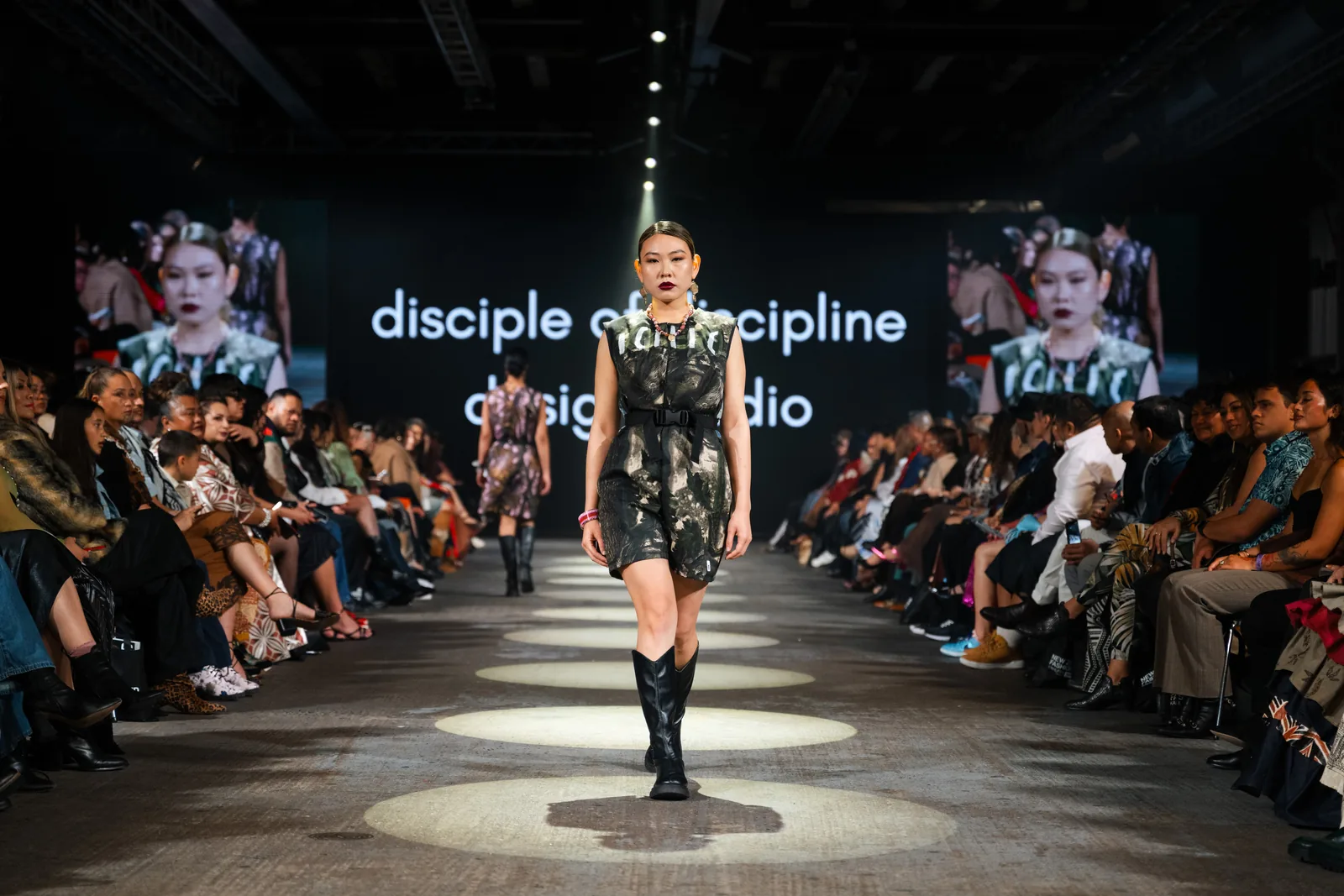 A dark print dress with a black belt and tall black boots on the runway